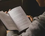 Młoda kobieta w szarej bluzie czyta książkę. Jej dłonie delikatnie trzymają otwarte strony.