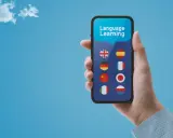 Dłoń trzyma smartfon z aplikacją do nauki języków. Wybór między Busuu czy Duolingo? Na ekranie flagi: UK, Hiszpanii, Niemiec, Francji, Chin, Japonii, Włoch, Polski.