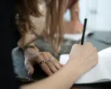 Dłoń z pierścionkami pisze w notatniku. Na długopisie widoczny jest napis "list oficjalny po angielsku".