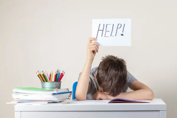 Chłopiec z kartką "HELP!" siedzi przy biurku ze stosem książek i kolorowych kredek.