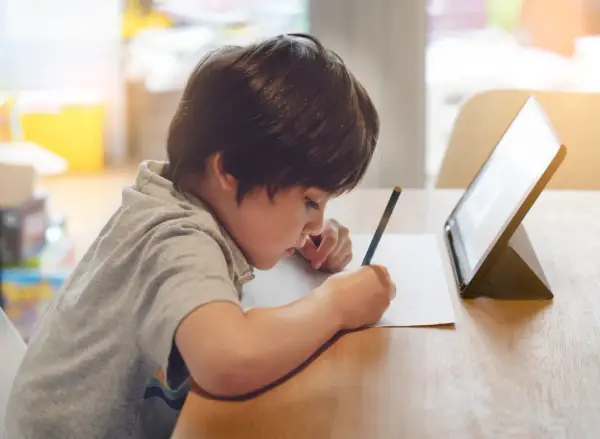 Chłopiec pisze ołówkiem na kartce, przed nim stoi tablet.